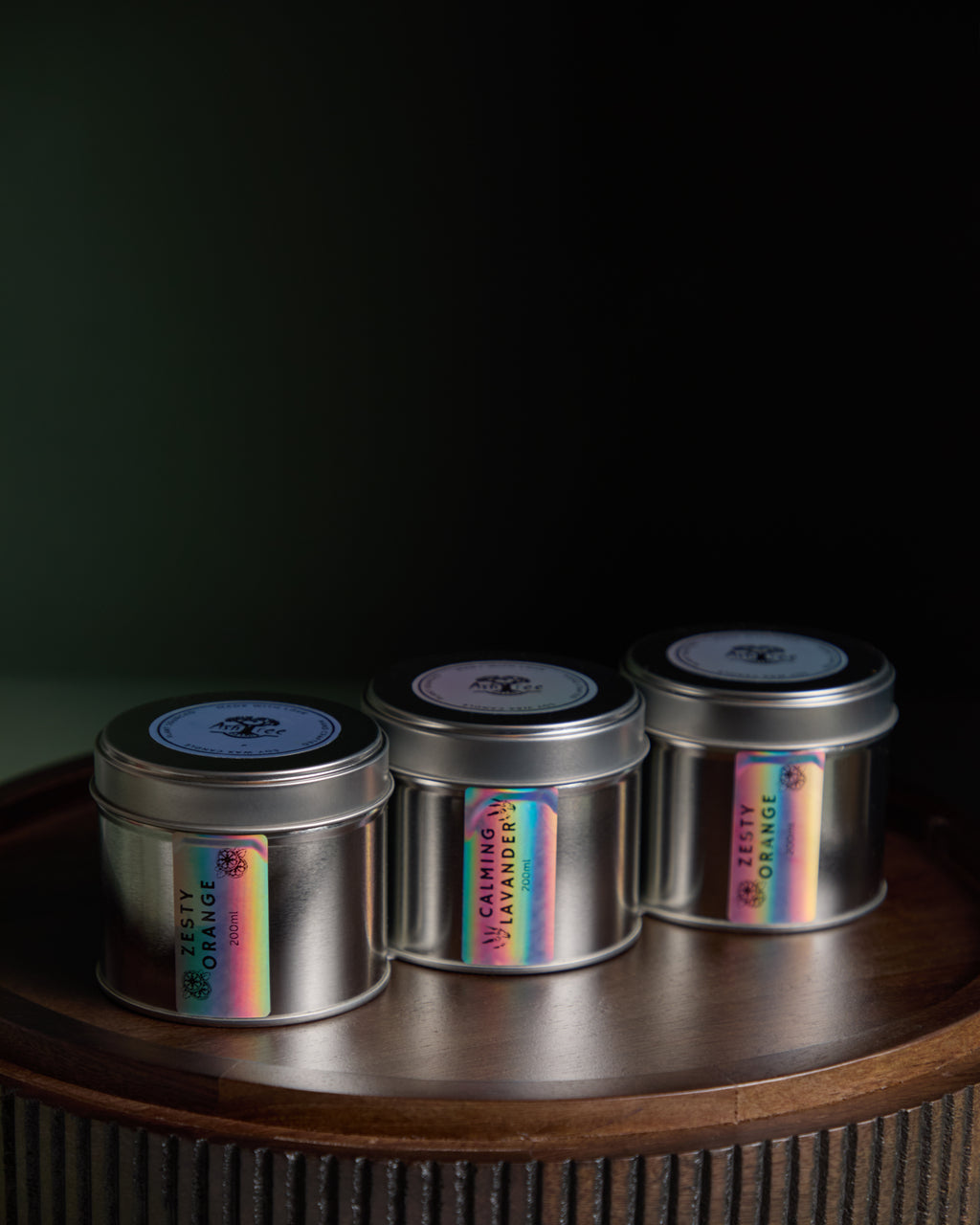 Three metal candles with holographic labels on a wooden surface against a dark background. A tin candle collection from the brand Ash Tree Candles 