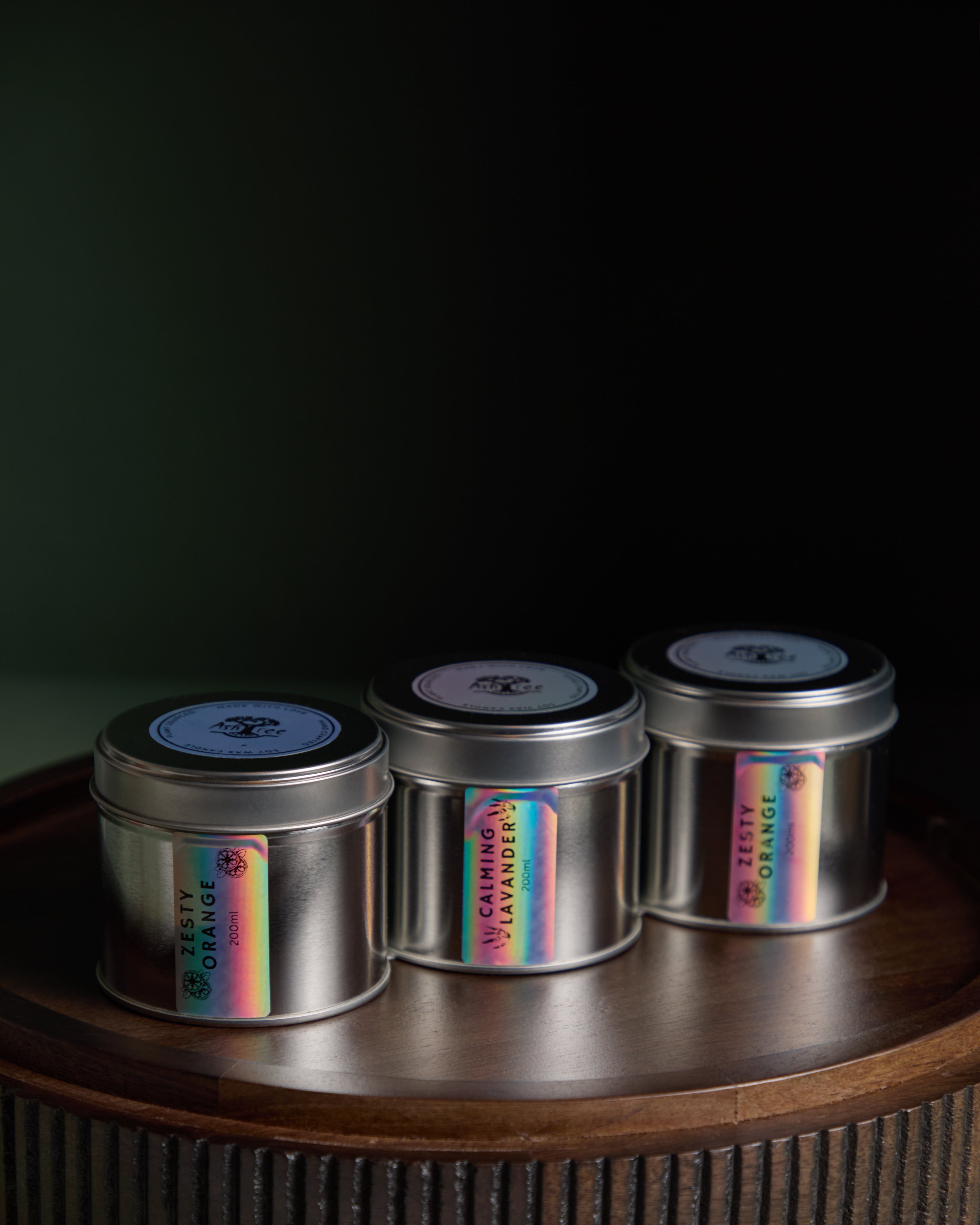 Three metal candles with holographic labels on a wooden surface against a dark background. A tin candle collection from the brand Ash Tree Candles 
